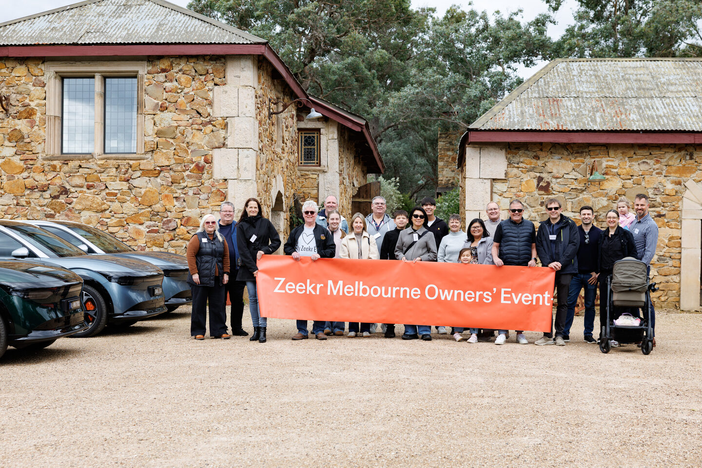ZEEKR owners and families posing with an orange event banner beside several ZEEKR vehicles and stone buildings during the Kinglake drive.